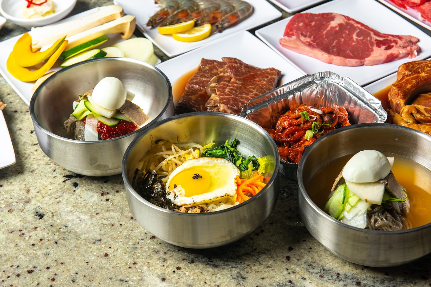Bibimbap and naengmyeon bowls beside plates of fresh meat